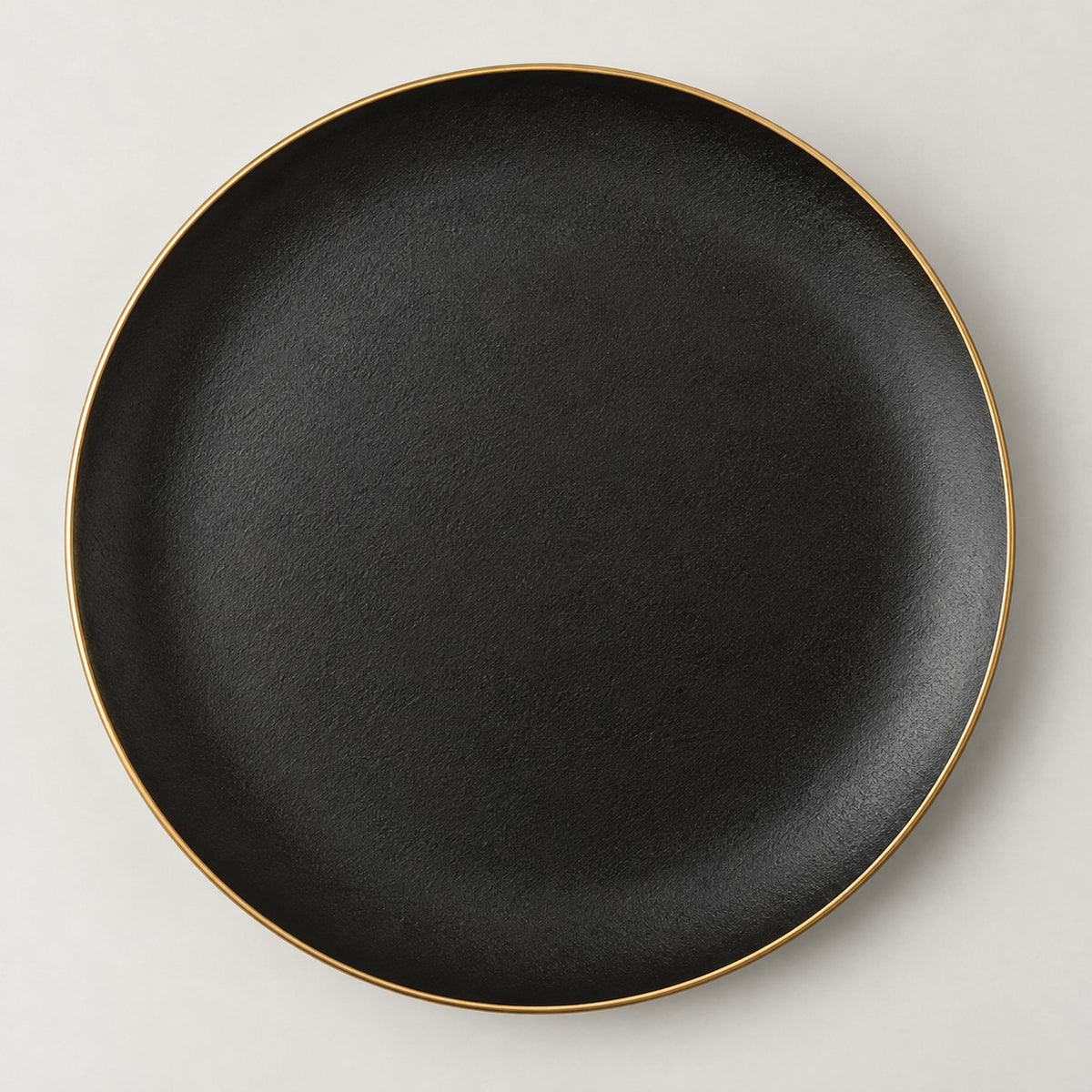 Single shallow black ceramic plate with a delicate gold rim centered on a light studio background.