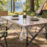 Clear disposable plastic tablecloth covers an outdoor camping table beneath picnic plates and a lantern.