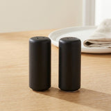 Two matte black cylindrical salt and pepper shakers standing side by side on a light wood tabletop, with a blurred white plate and folded cloth in the background.