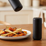 A matte black cylindrical salt and pepper grinder with a matching cylindrical container pouring seasoning over roasted vegetables on a white plate, set on a wooden table.