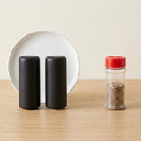 Two matte black cylindrical salt and pepper shakers stand on a light wood surface in front of a white plate, with a clear spice jar featuring a red lid and brown seasoning visible to the right.