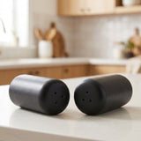 Two matte black cylindrical salt and pepper shakers resting on a white kitchen countertop with a blurred kitchen background.