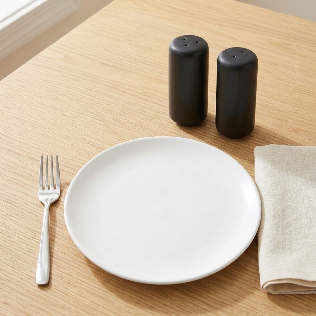 A minimalist dining table setting featuring a round white ceramic plate, a silver metal fork, a folded beige linen napkin, and two matte black cylindrical salt and pepper shakers on a light wood grain tabletop.