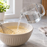A 2-cup clear glass measuring cup pours water into a mixing bowl on a kitchen counter.