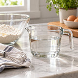 A clear glass 2-cup measuring cup holds liquid beside a bowl and whisk for home baking.