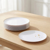 A stack of white ceramic plates with a thin gold rim sits on a light wood table next to one empty plate with a few crumbs, in a softly lit indoor setting.