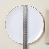 A white round plate with a thin gold rim, shown from above on a light beige textured background with a metal ruler placed vertically along the center for scale.