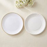 Two white ceramic plates on a light beige textured background, with one plate featuring a thin gold rim and the other featuring a fluted edge, styled with small white flowers at the top.