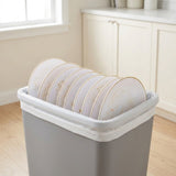 A gray rectangular trash can with a white plastic liner and a white plate-holding loop holding a stack of white plates with gold rims, set in a bright kitchen environment.