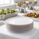 A stack of white dinner plates with a thin gold rim sits on a white tablecloth in a buffet setting, with salads and chafing dishes blurred in the background.