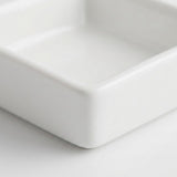 A close-up of a square white ceramic dish or small tray, showing the corner and smooth glossy surface on a white background.
