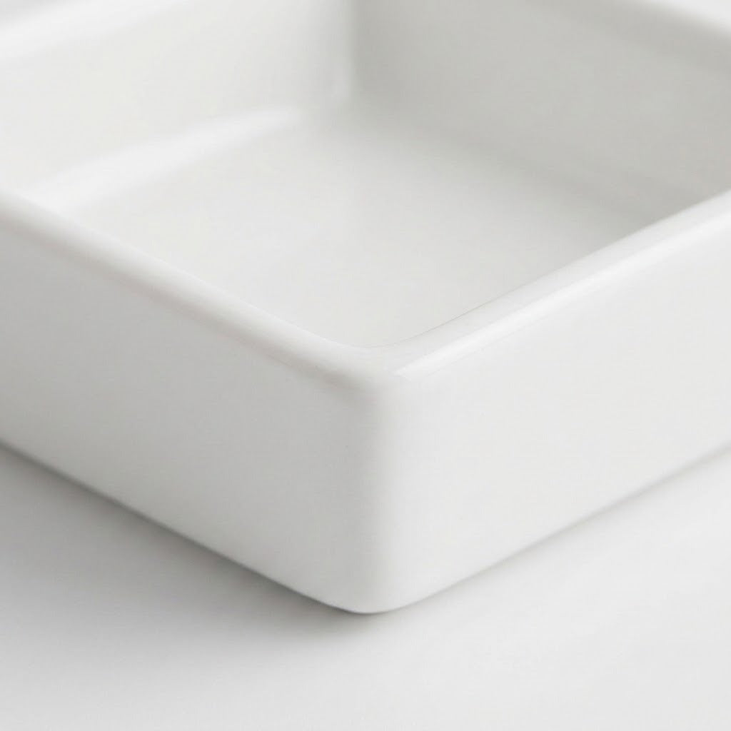A close-up of a square white ceramic dish or small tray, showing the corner and smooth glossy surface on a white background.
