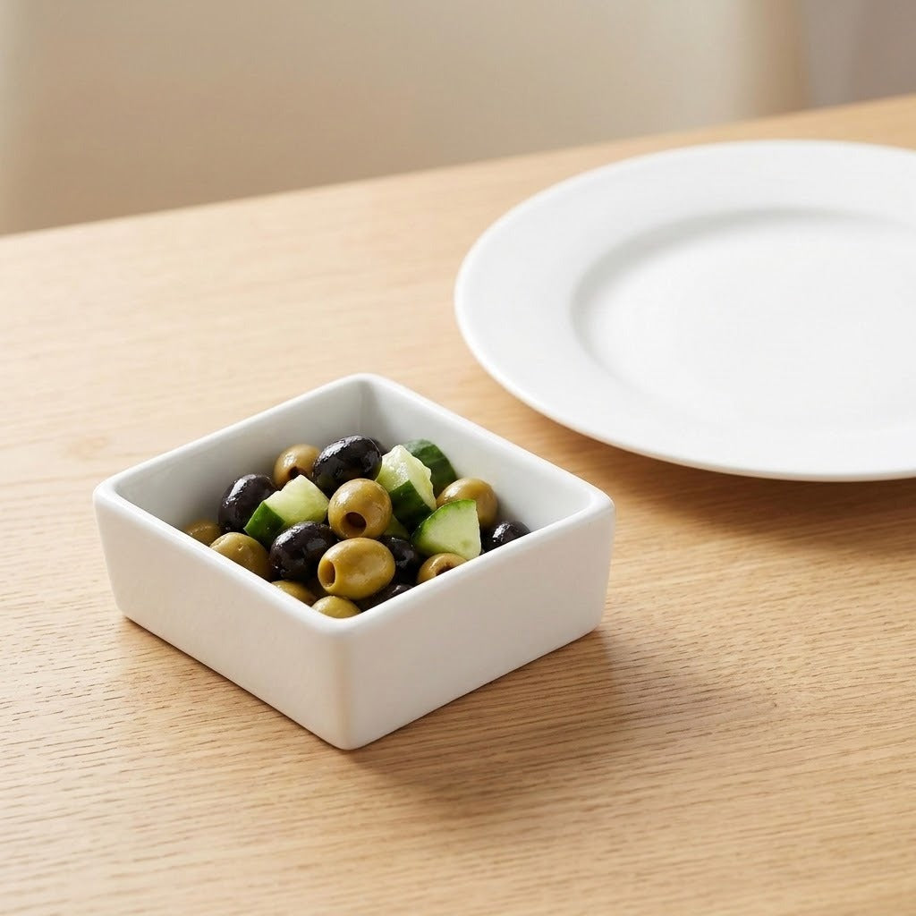A small square white ceramic bowl filled with sliced black olives, green olives, and cucumber, placed on a light wood table next to a white plate.