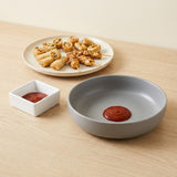 A gray ceramic serving bowl with a small red sauce dollop inside, shown alongside a white square dipping dish with ketchup and a plate of food on a light wood tabletop.