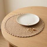 A round woven placemat with a scalloped edge in a natural tan color, styled on a light wood table with a small white ceramic plate on top.