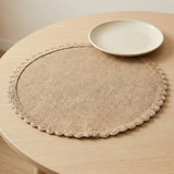 A round beige woven placemat with a scalloped edge sits on a light wood table, with a small off-white plate placed on top.