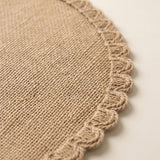 Close-up of a round woven placemat or rug in natural tan burlap-like fabric with a braided scalloped edge, shown on a light neutral background.