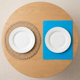 Top-down view of a round wooden table set with two white plates: one on a beige woven placemat and one on a bright blue square placemat.