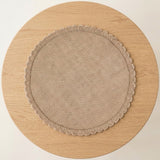 A round beige fabric placemat with a scalloped edge, displayed flat on a round light wood table against a plain off-white background.