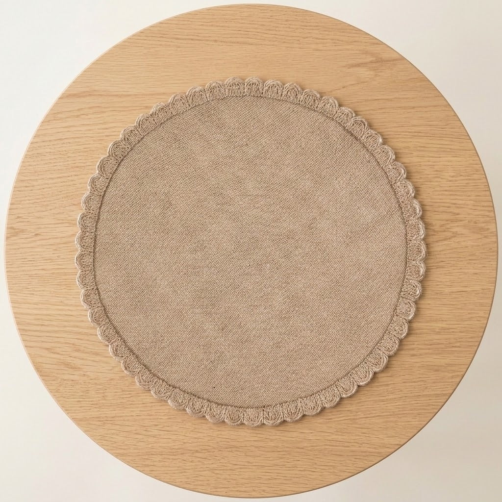 A round beige fabric placemat with a scalloped edge, displayed flat on a round light wood table against a plain off-white background.
