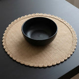A matte black ceramic bowl placed on a round beige woven placemat with a scalloped edge, set on a dark wooden table.