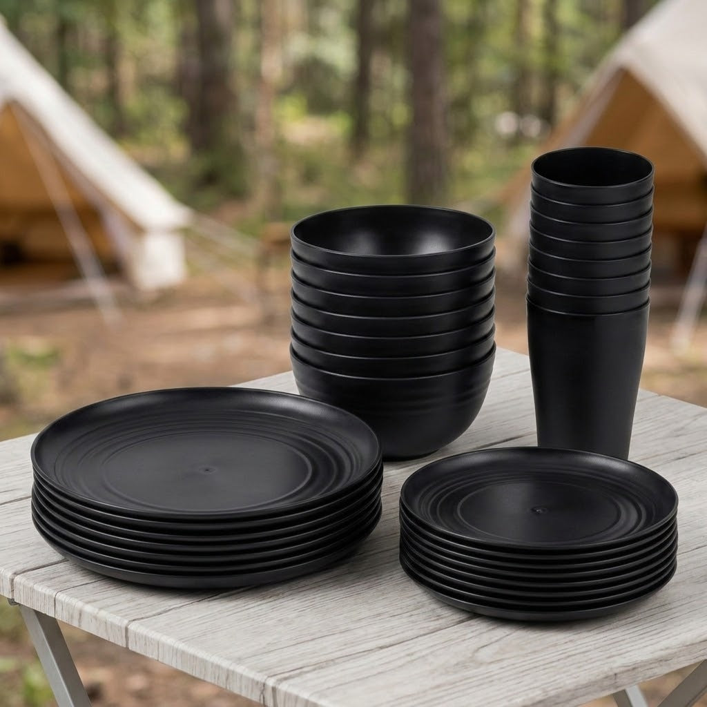 A set of matte black outdoor tableware including plates, bowls, and cups, arranged on a light-colored folding table with a blurred forest and tent background.