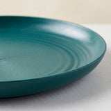 A close-up of a round teal ceramic plate with concentric ring details, resting on a light gray surface against a warm beige background.