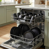 A stainless steel dishwasher with both racks pulled out, filled with black plates and bowls, set in a modern kitchen with light green cabinets and a white marble countertop.