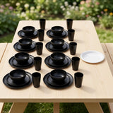 A set of matte black dinnerware, including bowls, plates, cups, and a white plate, arranged on a light wood picnic table outdoors with a blurred green garden background.