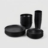 A set of matte black dinnerware, including plates, bowls, and cups, neatly stacked in three groups on a light gray background.
