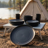 A set of matte black camping tableware, including plates and cups, arranged on a folding camp table outdoors with a tent and lake in the background.