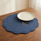 A scalloped dark blue quilted placemat on a wooden table, styled with a light beige ceramic plate and a few crumbs.