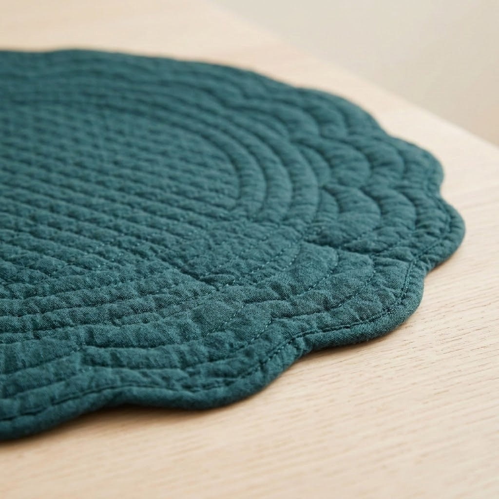 A close-up of a teal green quilted placemat with a scalloped edge, resting on a light-colored wooden tabletop.