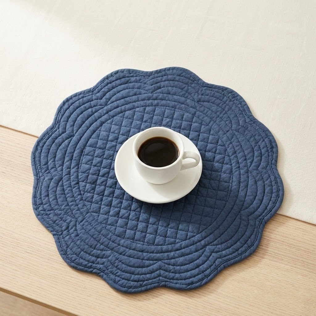 A scalloped blue quilted placemat on a light wood table, styled with a white cup of coffee on a saucer in the center.