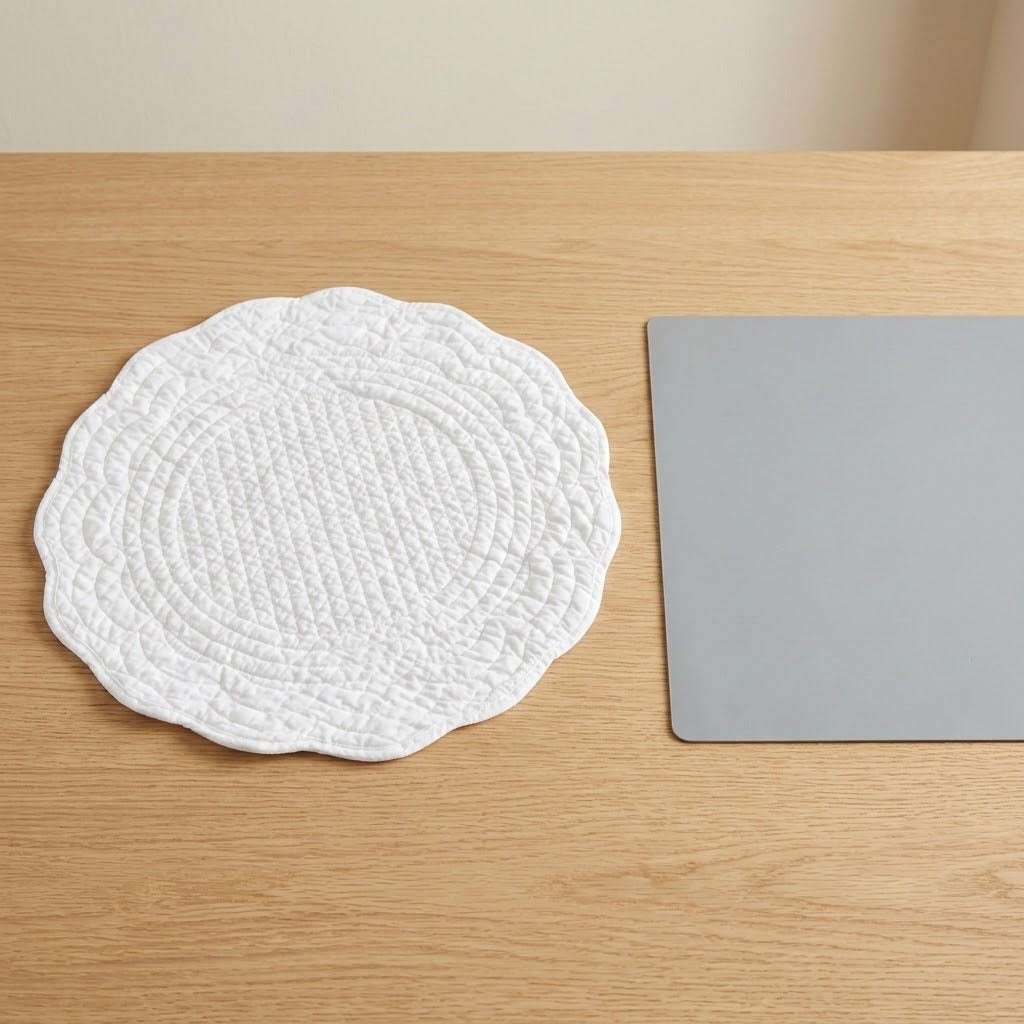 A round white quilted placemat with a scalloped edge sits on a light wood table next to a gray rectangular board.