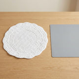 A round white quilted placemat with a scalloped edge sits on a light wood table next to a gray rectangular board.