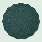 A round quilted placemat with a scalloped edge in a dark teal green color, shown flat against a light mint green background.