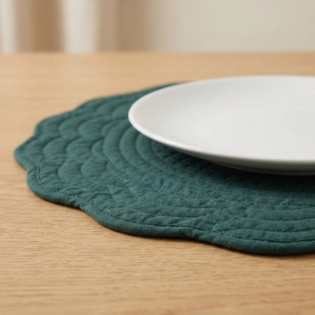 A dark teal quilted placemat with a scalloped edge on a light wood table, with a white ceramic plate partially resting on top.