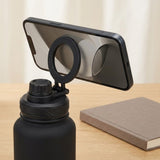 A black phone mount with a circular ring and clip, shown attached to a matte black water bottle on a light wood table, with a smartphone resting in the mount and a neutral book in the background.