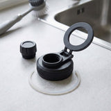 A black plastic water bottle lid with an open hinged flip-up spout and a separate cap, placed on a light countertop near a sink. A clear silicone seal is visible beneath the base.