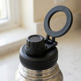 Close-up of a stainless steel water bottle with a black flip-top lid and a hinged carrying loop, photographed near a window.