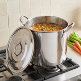 A large stainless steel stockpot with its lid leaning against it, filled with a vegetable soup on a gas stovetop in a kitchen. A bunch of carrots and celery sits on the counter beside the pot.