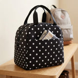 A black and white polka dot lunch bag with dual top handles sits on a light wood table, with a gray backpack blurred in the background.