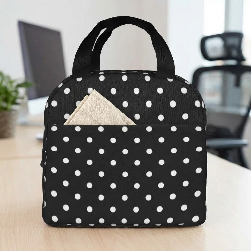 A black insulated lunch bag with white polka dots sits on a light wood desk, with a beige napkin tucked into the front pocket. The background shows a softly blurred office setting with a computer monitor and chair.