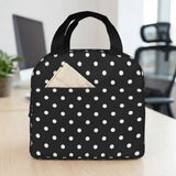 A black insulated lunch bag with white polka dots sits on a light wood desk, with a beige napkin tucked into the front pocket. The background shows a softly blurred office setting with a computer monitor and chair.