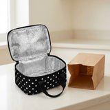 A black insulated lunch bag with white polka dots and a silver reflective lining, shown open on a light countertop next to a brown paper bag.