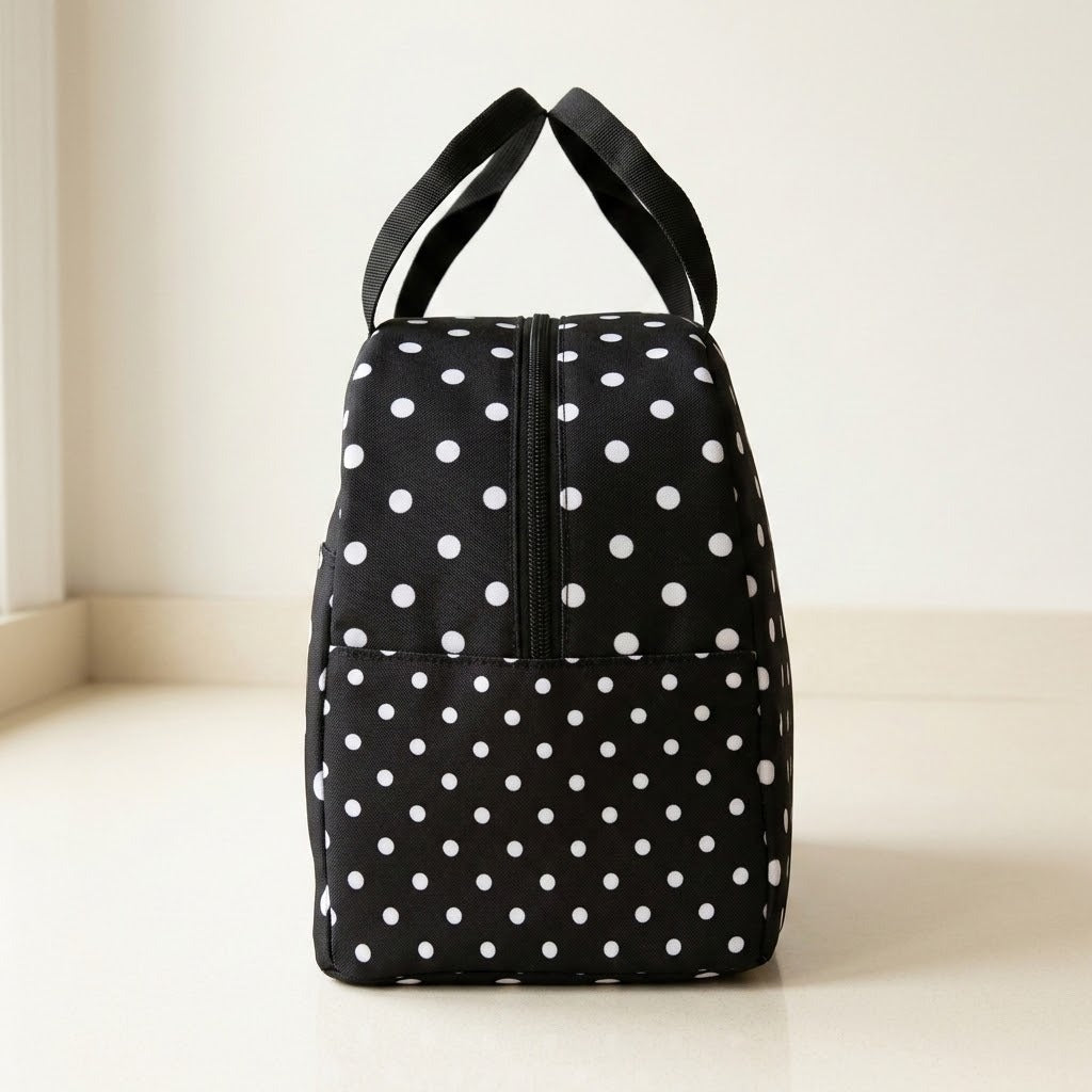 A black soft-sided bag with white polka dots and two black shoulder straps, standing upright on a light beige floor against a cream wall.