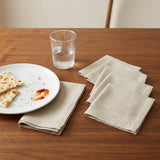 A set of light beige linen napkins arranged on a wooden table, with one folded napkin under a white plate with crumbs and a piece of bread, and another folded napkin beside a clear glass of water.