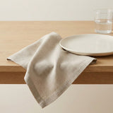 A light beige linen napkin draped over a light wood table next to a white ceramic plate and a glass of water.