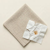 A beige textured fabric square, likely a napkin or small towel, shown on a light cream background with a crumpled white paper towel featuring a brown stain placed on top.